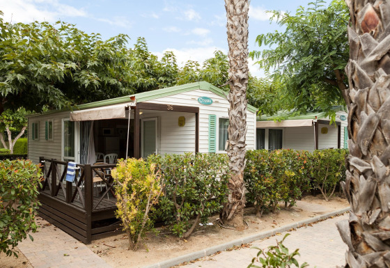 Mobilhjem Camping Cambrils Playa BUNGALOW OHARA med terrasse og omgivet af grønne buske og palmetræer.