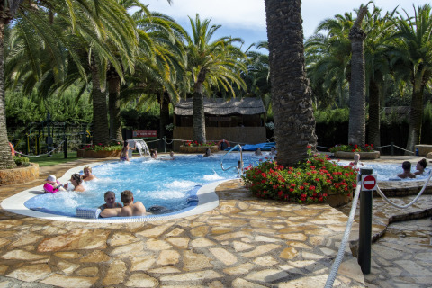 Udendørs poolområde med palmer og mennesker, set på et glamping-ophold under solrige himmelstrøg.