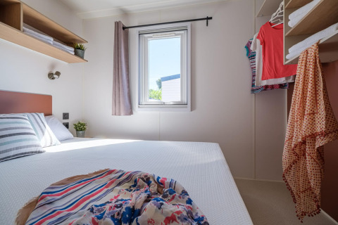 Helles, kompaktes Schlafzimmer in einem Mobilheim mit Doppelbett, Kleidung und Fenster mit Vorhang.