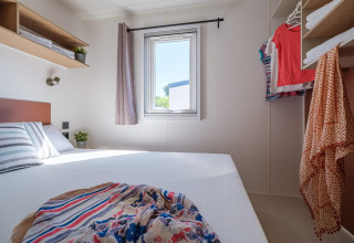 Luminoso dormitorio compacto en casa móvil con cama doble, ropa colgada y ventana soleada.