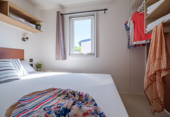 Lichte, compacte slaapkamer in een mobilhome met tweepersoonsbed, kledingrek en raam met gordijn.
