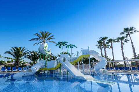 Waterspeelplaats met glijbanen en palmbomen bij een glamping accommodatie onder een heldere blauwe lucht.
