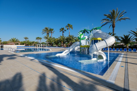 Piscina exterior con toboganes y palmeras en un alojamiento glamping bajo un cielo azul claro.