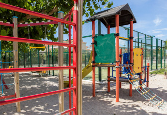 Area giochi con struttura per arrampicata e scivolo, sotto alberi, in un glamping con campi sportivi.