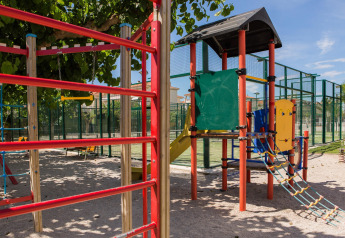 Speelplaats met klimrek en glijbaan onder bomen bij een glampinglocatie met sportvelden.