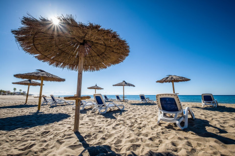Strand med liggestole og stråparasoller under en klar blå himmel ved glamping-overnatning.