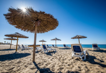 Strand med liggestole og stråparasoller under en klar blå himmel ved glamping-overnatning.