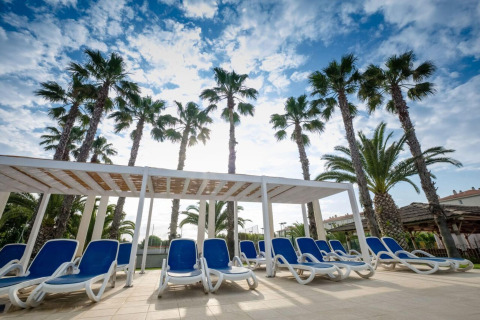 Solrige blå solsenge under pergola, omgivet af palmer i Camping Estival Vendrell Platja Grand Safari Glamping, Spanien.