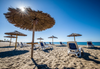 Plage de sable avec parasols en paille et transats sous ciel bleu à Camping Estival Vendrell Platja, Espagne