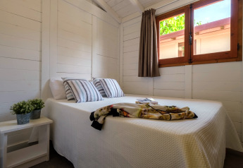 Chambre lumineuse avec lit double, coussins rayés, fenêtre et plante au Camping Estival Vendrell Platja, Espagne.