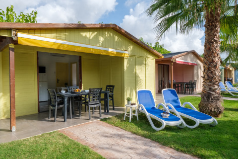 Udendørsbillede af en gul bungalow med terrasse, solsenge og palme på Camping Estival Vendrell Platja i Spanien.