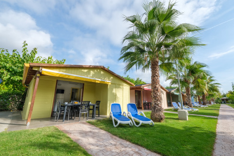 Solrige bungalower på Camping Estival Vendrell Platja med palmer og liggestole på grøn græsplæne.