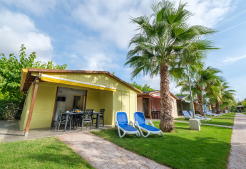 Kleurrijke bungalows op Camping Estival Vendrell Platja met palmbomen, ligbedden en groen gras.