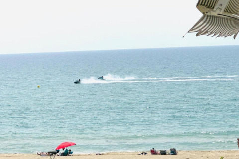 Vista desde una cabaña hacia la playa con personas bajo una sombrilla roja y motos acuáticas en el mar.