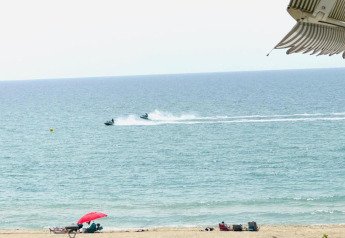 Uitzicht vanuit een lodge op een strand met mensen onder een rode parasol en jetski’s op zee.