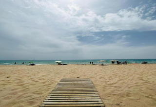 Strandfoto taget ved Camping Estival Vendrell Platja i Spanien med træsti, sand, parasoller og hav.