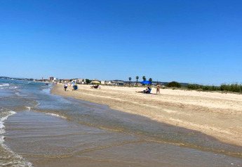 Scène de plage paisible avec quelques personnes sur le sable et des bâtiments au loin sous un ciel bleu clair.