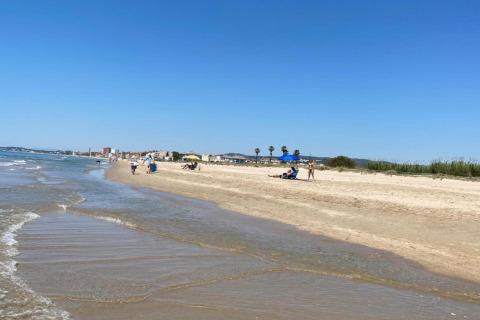 Udsigt over en fredelig strand med få mennesker, solsenge og bybygninger i baggrunden under en blå himmel.