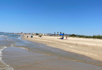 Rustig zandstrand met enkele mensen, zonneschermen en gebouwen op de achtergrond onder een heldere blauwe lucht.