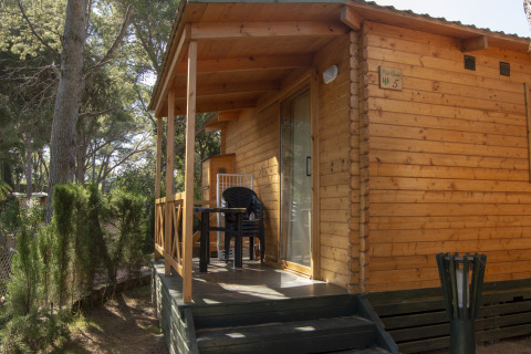 Træhytte glamping indkvartering i skovområde med terrasse og plastikmøbler under træerne.