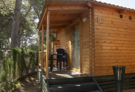 Træhytte glamping indkvartering i skovområde med terrasse og plastikmøbler under træerne.