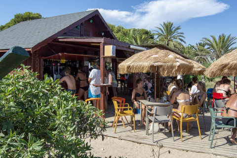 Persone sedute sotto ombrelloni di paglia in un glamping, vicino al bar in legno all’aperto sulla spiaggia.