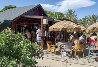 Folk nyder drinks under stråparasoller ved et glamping-sted med en udendørs bar og masser af sol.