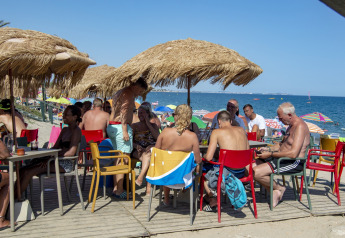 Persone che si rilassano sotto ombrelloni di paglia in una sistemazione glamping con sedie colorate al mare.