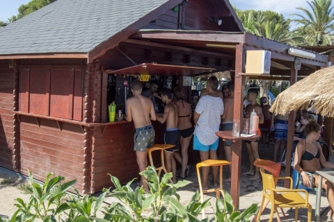 Folk i badetøj samles ved en strandbar i træ på et glampingsted omgivet af palmer og planter.