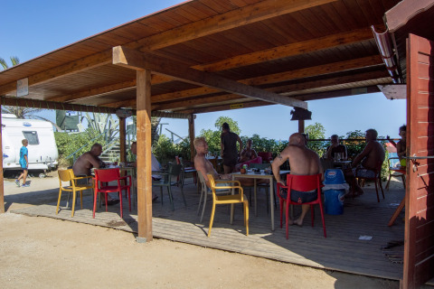 Personas sentadas bajo un pabellón de madera en un alojamiento glamping, rodeados de naturaleza y caravanas.