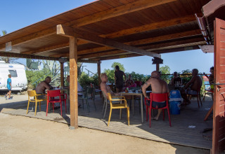 Personas sentadas bajo un pabellón de madera en un alojamiento glamping, rodeados de naturaleza y caravanas.