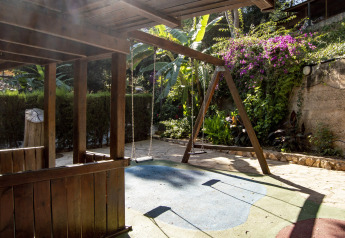 Parco giochi con altalena e casetta di legno in una struttura glamping circondata da giardino rigoglioso.