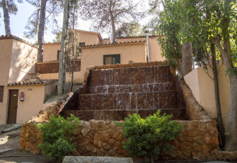 Vista esterna di un alloggio glamping con cascata, alberi e case dai toni terracotta tutto intorno.