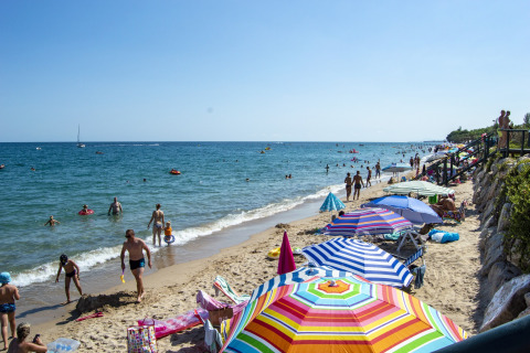 Strandscene med farverige parasoller, folk der bader og nyder solen ved et glamping-ophold.