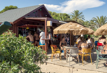 Bar de plage en plein air dans un hébergement glamping, clients en maillot sous des parasols en paille.