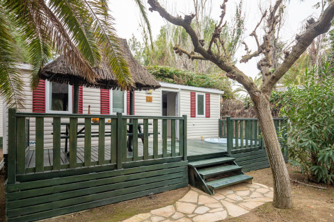 Et mobilhome med grøn veranda og stråtag i Spanien, omgivet af træer og planter, set fra haven.