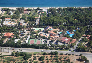 Luftfoto af glamping sted med swimmingpool, tennisbaner, parkeringsplads og strand i baggrunden.