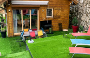 Zona esterna di uno chalet in legno con erba artificiale, sedie colorate e lettini vicino a un muro di pietra.