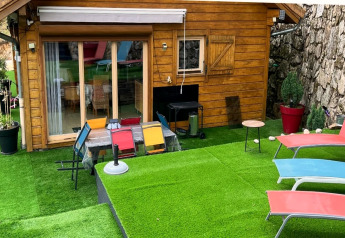 Zona esterna di uno chalet in legno con erba artificiale, sedie colorate e lettini vicino a un muro di pietra.