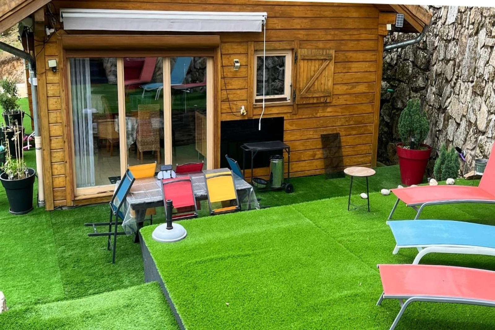 Buitengebied van een houten chalet met kunstgras, kleurrijke stoelen en ligbedden naast een stenen muur.