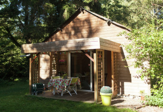 Cabaña de madera con porche y mesa de jardín rodeada de naturaleza en Maison de vacances - LE PONCHEL, Francia.