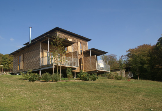 Moderne houten lodge op een groene helling, omgeven door natuur en bomen onder een blauwe hemel.