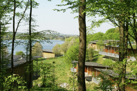 Uitzicht op een lodge in een bosrijk landschap met een meer op de achtergrond tijdens een zonnige dag.