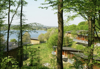 Uitzicht op een lodge in een bosrijk landschap met een meer op de achtergrond tijdens een zonnige dag.