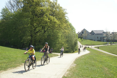 Mensen fietsen en wandelen op een pad bij een lodge, tussen groene bomen en moderne gebouwen.