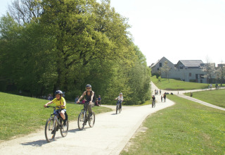 Mensen fietsen en wandelen op een pad bij een lodge, tussen groene bomen en moderne gebouwen.