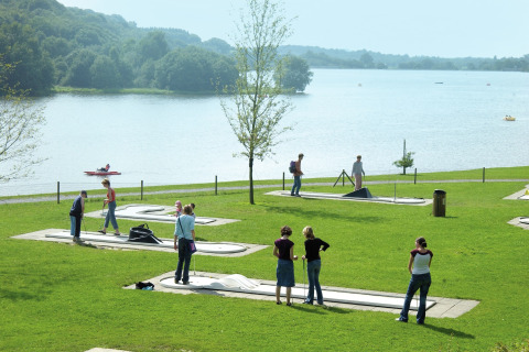 Mensen spelen minigolf op een grasveld bij een meer met kajaks aan een lodge in een groene omgeving.