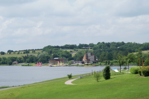 Foto van een lodge aan een meer, met zeilbootjes, groene velden en heuvels onder een bewolkte lucht.