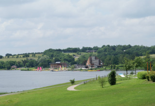 Foto van een lodge aan een meer, met zeilbootjes, groene velden en heuvels onder een bewolkte lucht.