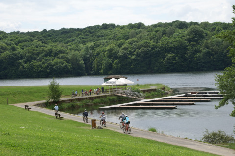 Fietsers rijden langs een meer met drijvende pontons en groene heuvels op de achtergrond bij een lodge.